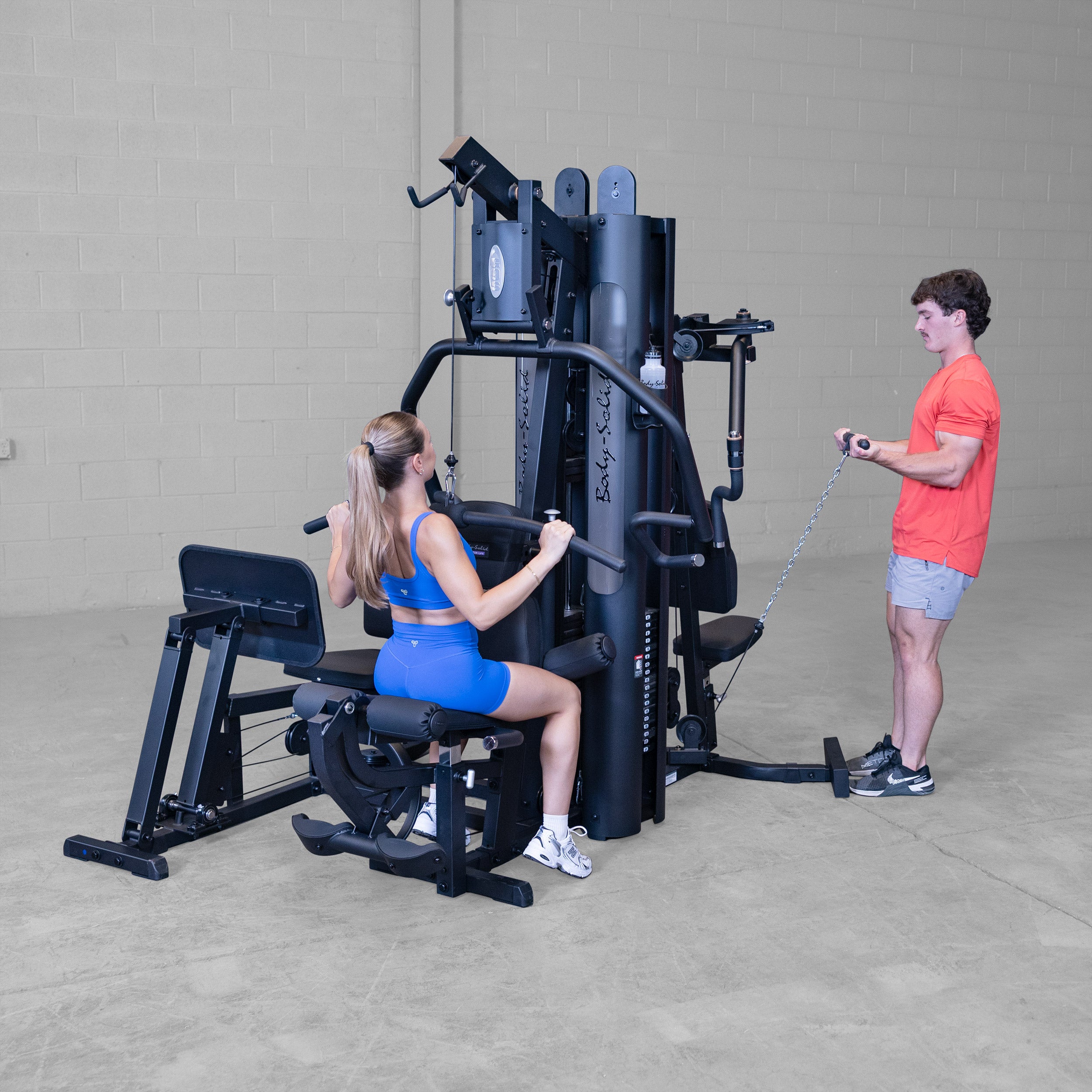 Two people training simultaneously on the Body-Solid G9B dual stack multi station home gym performing lat pulldown and standing biceps curl exercises