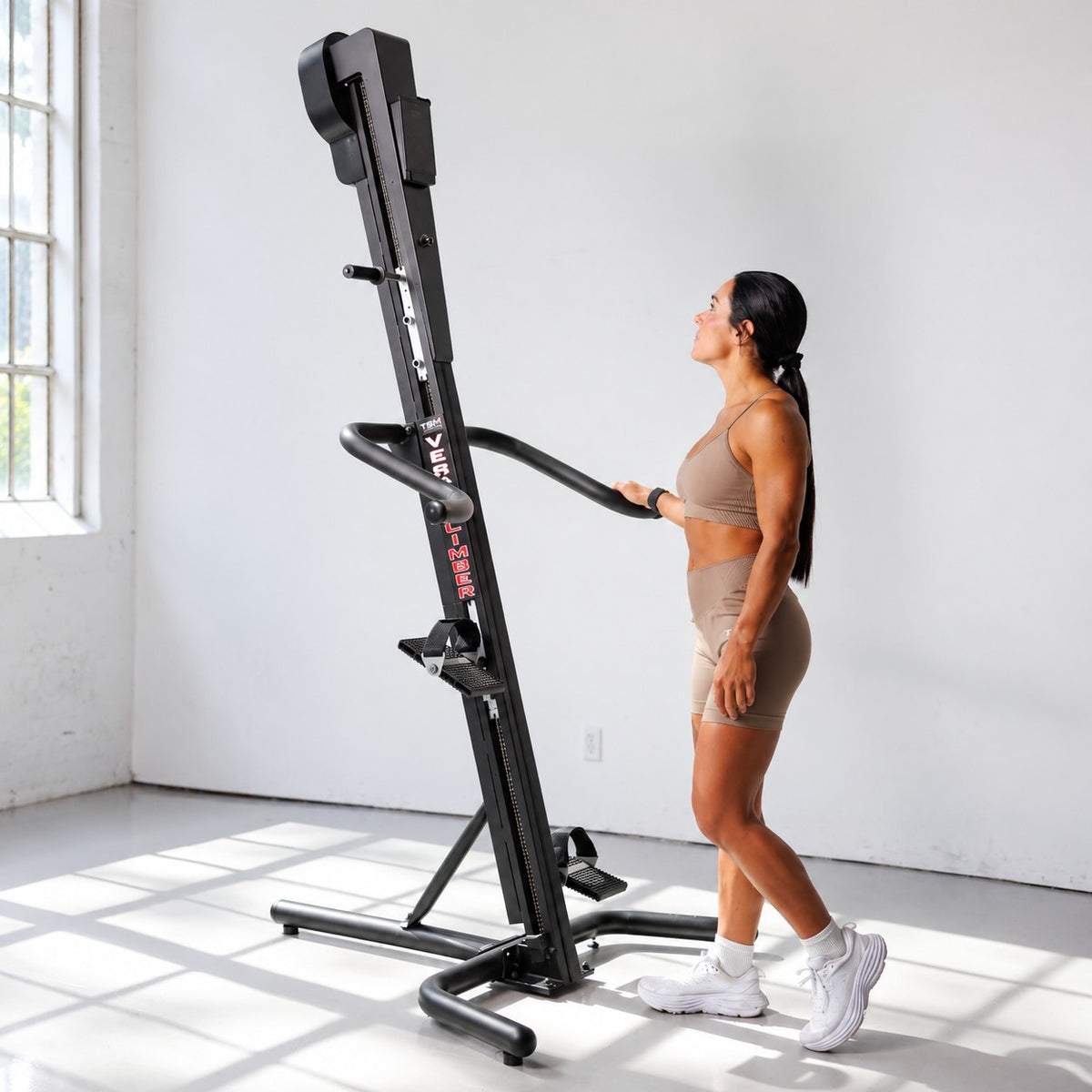 Woman standing beside VersaClimber TS-Magnetic vertical climber with standard base and holding the handlebar in a bright gym