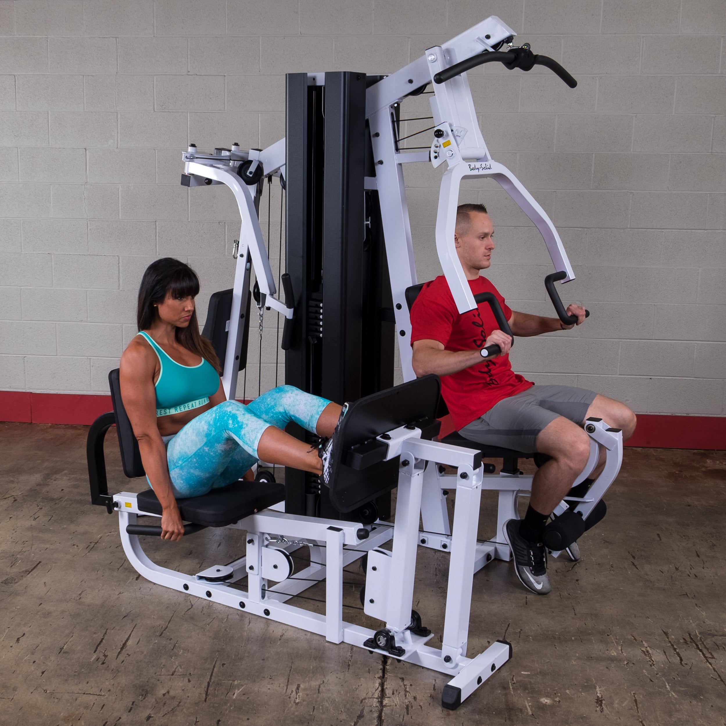 Two users training simultaneously on the Body-Solid EXM3000LPS dual stack multi-station gym performing chest press and leg press exercises