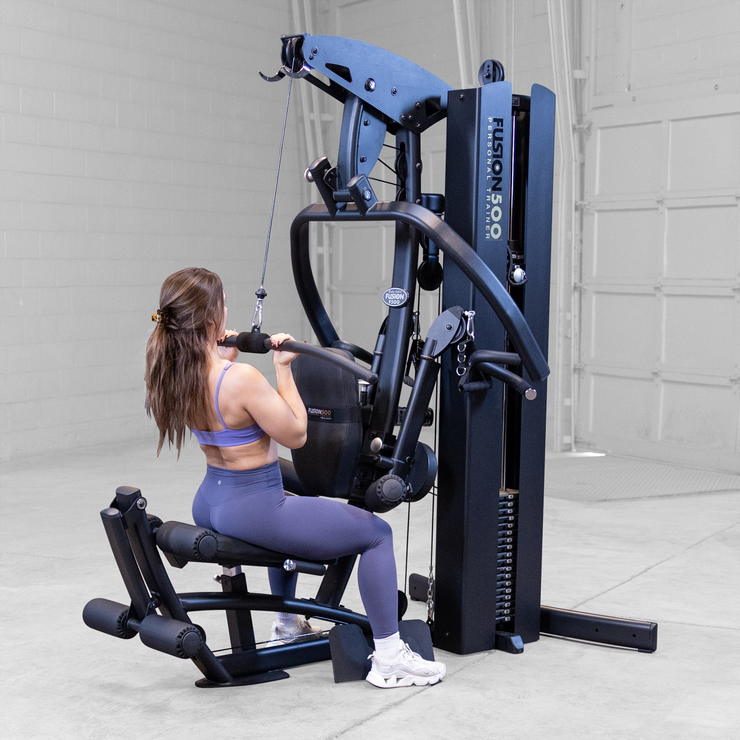 Person performing a lat pulldown on the Body-Solid Fusion 500B Personal Trainer using the high pulley and padded lat bar