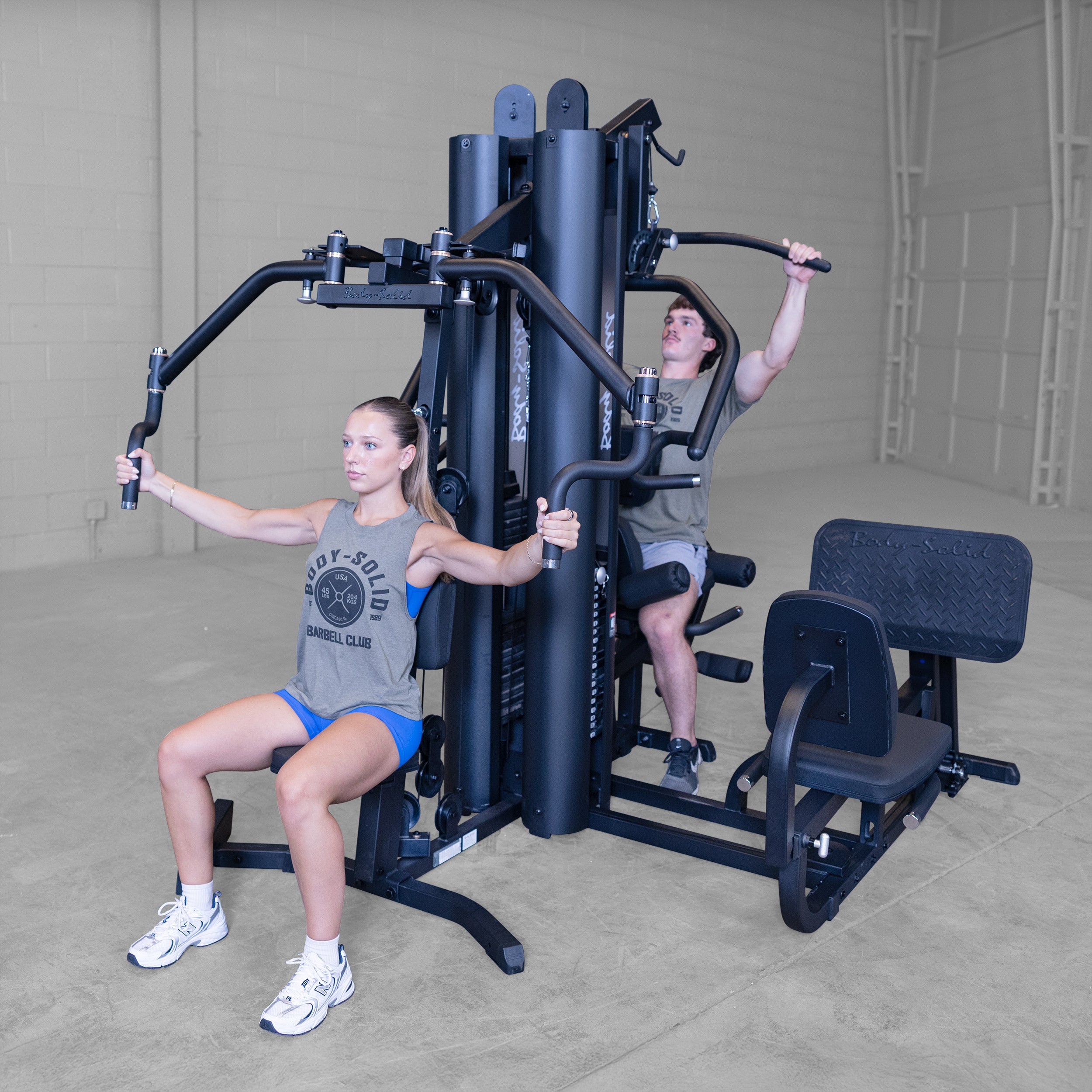 Two people performing chest press and lat pulldown exercises simultaneously on the Body-Solid G9B dual stack multi station home gym